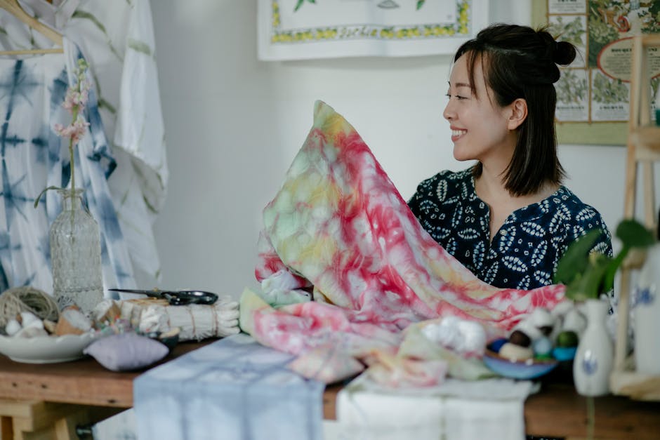 Positive Asian female dressmaker sitting at table with painted fabric creating handmade cloth in workshop