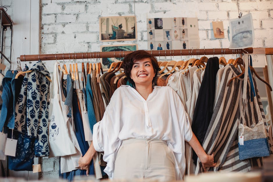 Satisfied young Asian female shopaholic standing near rack with various stylish clothes in fashion boutique and smiling